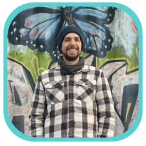 Brock Horning - Senior Copywriter - A white, bearded, smiling man stands in front of a graffitied metal container. He wears a blue beanie and chequered shirt.